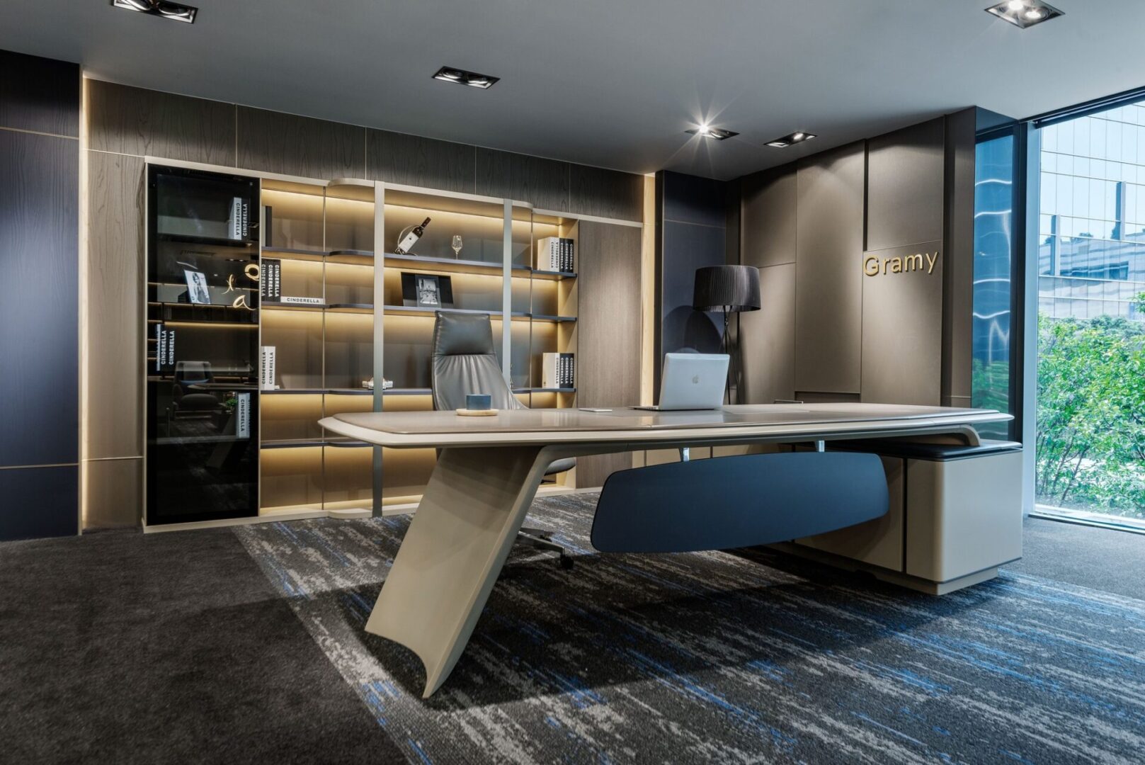 Modern office with a sleek desk, leather chair, and bookshelf. Large window with city view and "Gramy" logo on the wall. Neutral color palette and contemporary design.