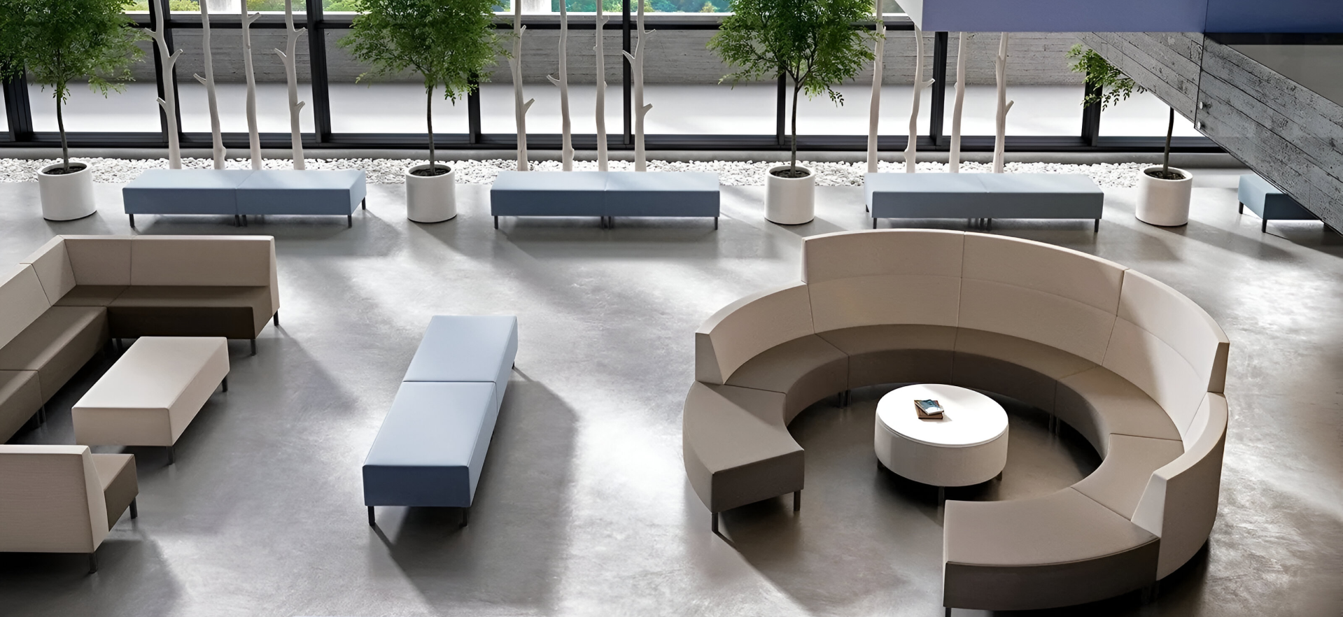 Modern lobby with circular and rectangular seating, a round table, potted trees, and large windows overlooking greenery.