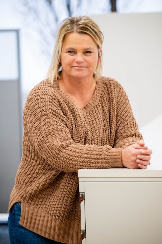 Woman in brown sweater, leaning on desk.